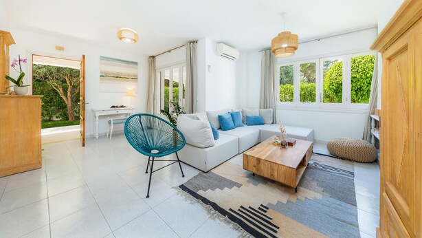 Una sala de estar moderna con un sofá blanco, una silla azul, una mesa de centro de madera y grandes ventanales con follaje exterior.