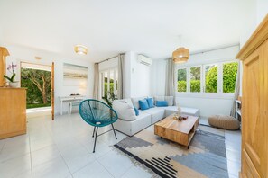 Uma sala de estar moderna com um sofá branco, uma poltrona azul, mesa de centro de madeira e grandes janelas com vegetação lá fora.