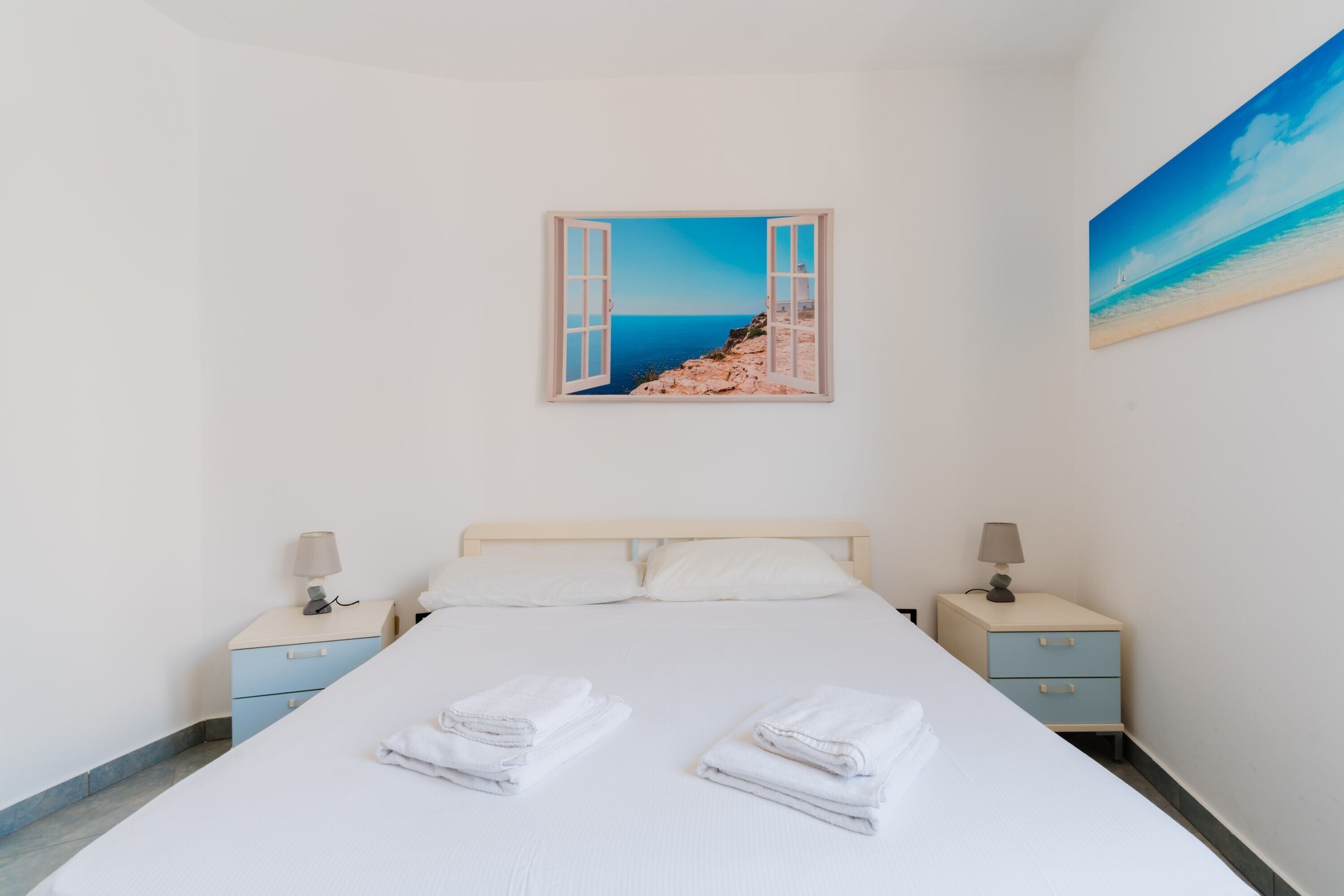 Ein Schlafzimmer mit einem Bett, zwei Nachttischen, einem Strandbild und Blick aufs Meer.