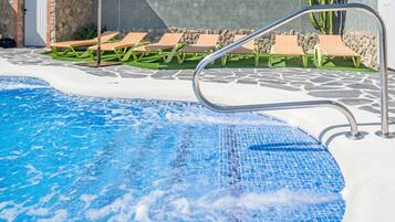 Une piscine avec un bord carrelé, des chaises longues et un mur en pierre.