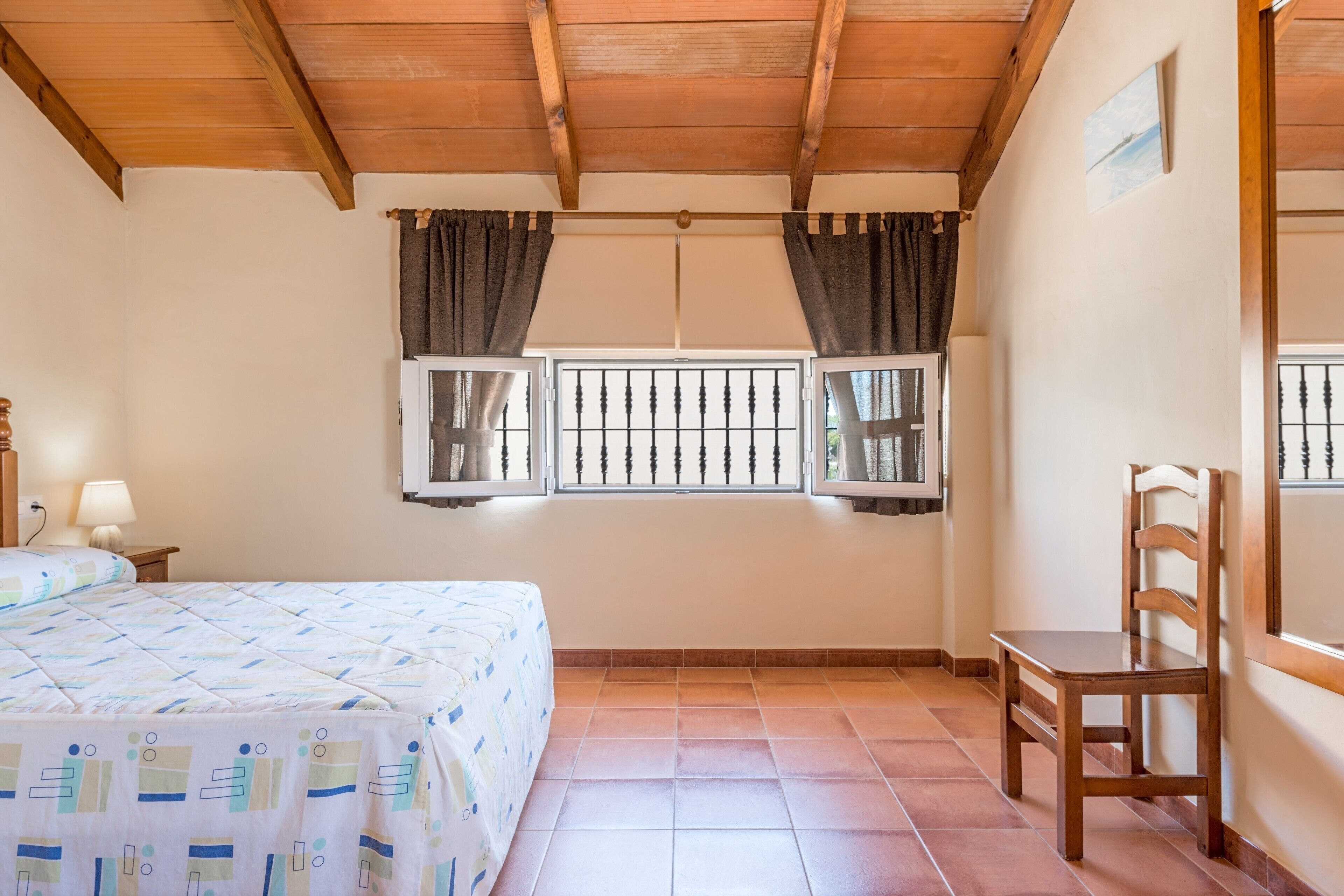 Un dormitorio con una cama, dos ventanas con cortinas, una silla de madera y una mesita de noche con lámpara.