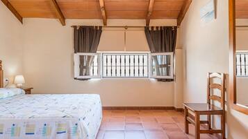 Un dormitorio con una cama, dos ventanas con cortinas, una silla de madera y una mesita de noche con lámpara.