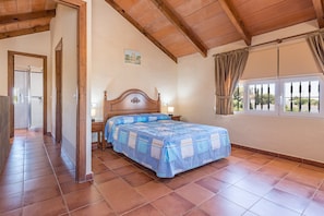 A bedroom with a wooden ceiling, a bed with a blue and white bedspread, a nightstand, a window with curtains, and a door leading to another room.