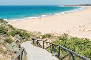 Ein hölzerner Steg führt zu einem Strand mit klarem, blauem Wasser und sandigem Ufer.