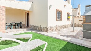 Ein weißes Gebäude mit rustikalem Steinunterbau, einer Terrasse mit grüner Kunstrasen und einem Tisch mit Stühlen.