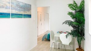 Una habitación moderna con pared blanca, una planta grande en maceta, una mesa blanca con dos sillas y una puerta que da a otra habitación.