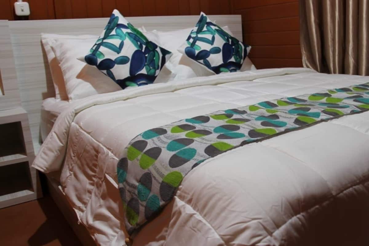 Un lit bien fait, recouvert d’une couette blanche, avec deux oreillers à motifs bleus et verts, et un plaid aux motifs verts et gris.