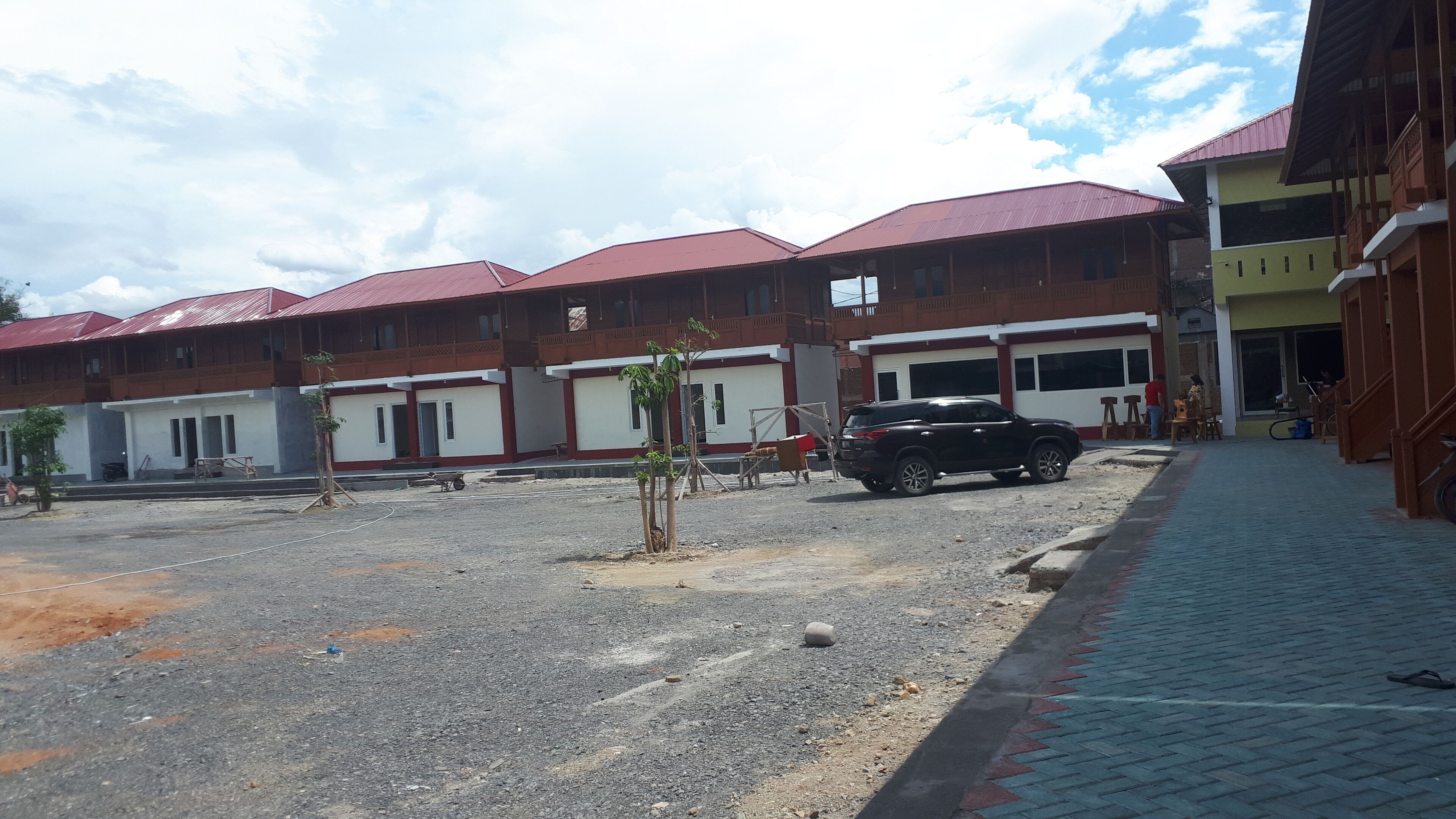 Un edificio con varias unidades, cada una con techo rojo y paredes blancas. Hay un área pavimentada al frente con un coche estacionado y algunos materiales de construcción.