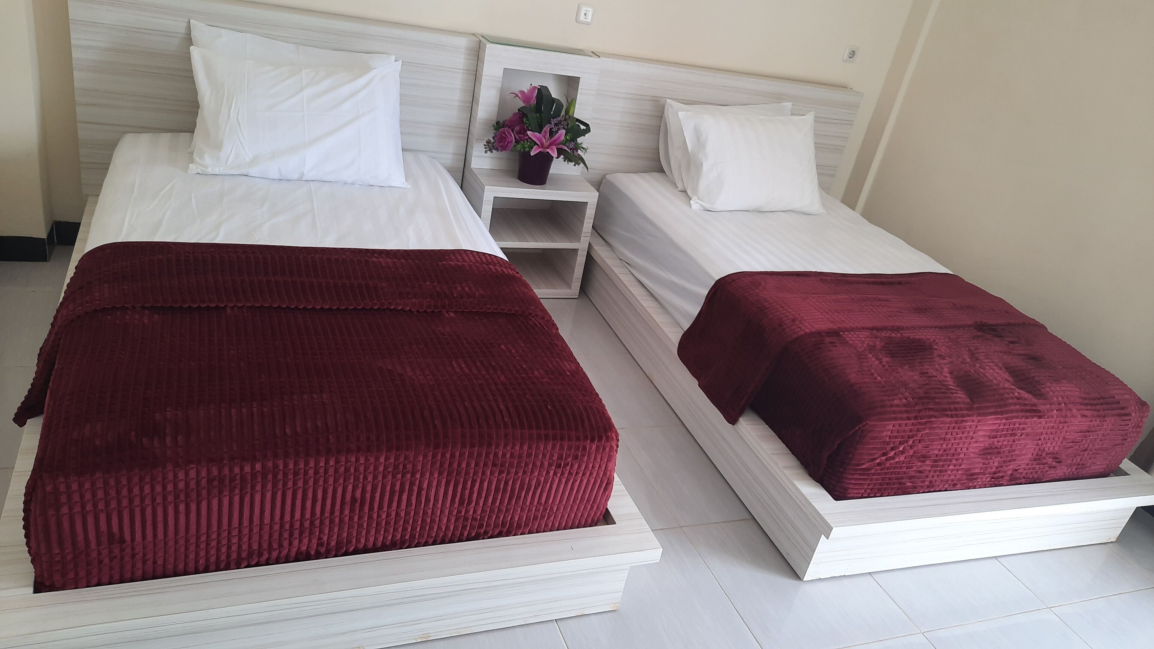 A hotel room with two beds, each with a white headboard and a red bedspread. There is a nightstand with a vase of flowers between the beds.