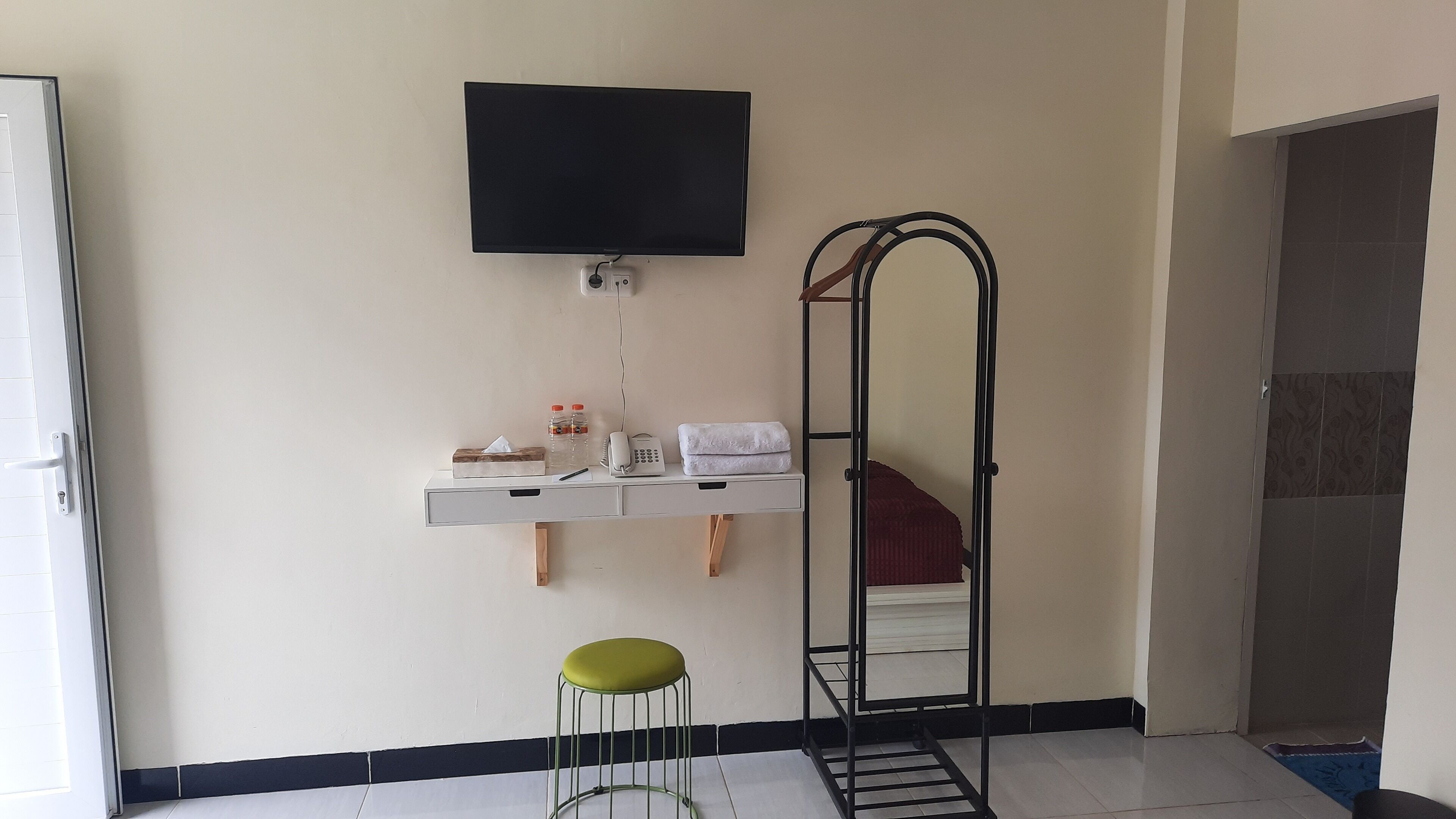 A room with a TV mounted on the wall, a mirror, a small table with toiletries, and a green stool.