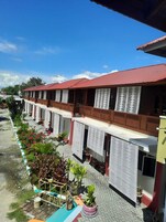 Una hilera de habitaciones de hotel con balcones, un paseo cubierto de baldosas y macetas con plantas.