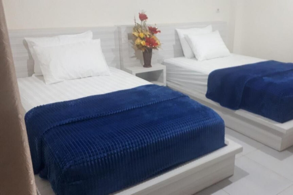 Dos camas con ropa de cama blanca y mantas azules, una mesita de noche con un florero y una pared blanca.