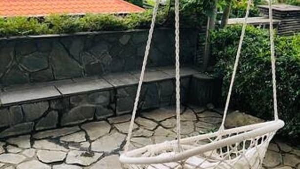 A wicker swing chair hanging from ropes, set against a stone-paved patio with a stone wall and greenery in the background. - Dot King Villa (Taipei)