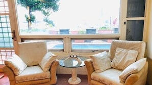 A room with two beige armchairs and a small glass table, a large window with a view of a pool and greenery outside. - Dot King Villa (Taipei)