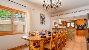A dining area with a wooden table set for six, surrounded by chairs, a chandelier, and a kitchen area with wooden cabinets and a stone countertop. - Air-conditioned Villa with Pool, Terrace and Wi-Fi (Sant Joan de Labritja)