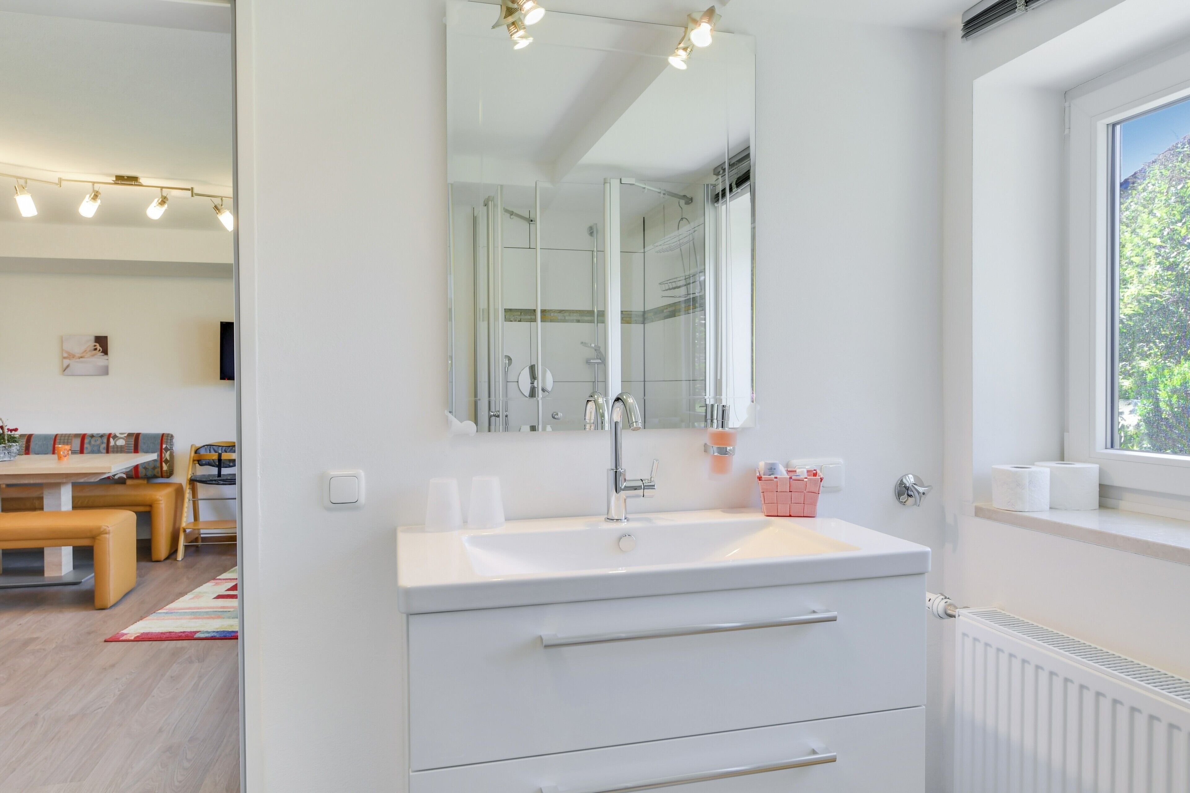 A modern bathroom with a large mirror, white sink, and a window with a view of greenery.