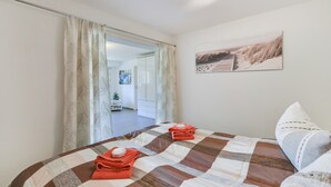 Un dormitorio con colcha a cuadros, dos toallas, un almohadón y un cuadro con motivos playeros.