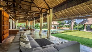 A covered outdoor seating area with a pool and lush greenery in the background.