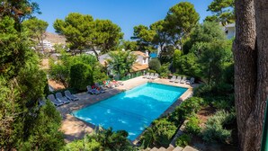 Une piscine entourée d’arbres et de bâtiments, avec des chaises longues à proximité.