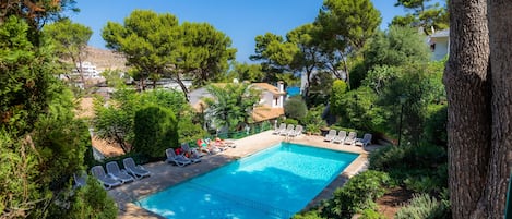 Une piscine entourée d’arbres et de bâtiments, avec des chaises longues à proximité.