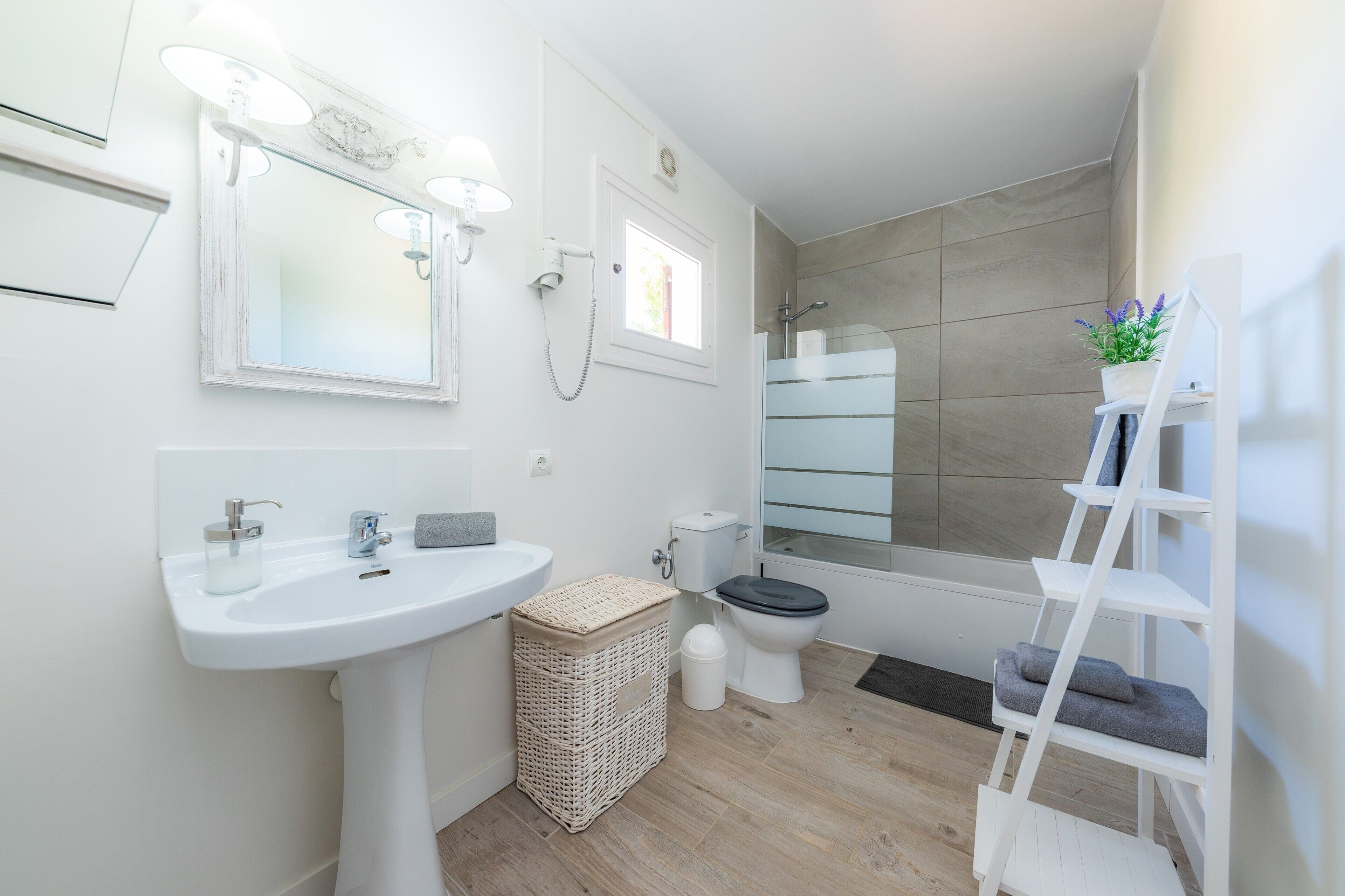 Une salle de bain moderne avec un lavabo blanc, un miroir et des toilettes, un petit panier en osier et une étagère blanche agrémentée d’une plante.