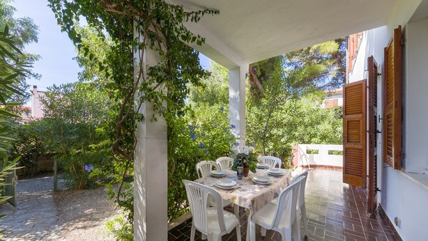 Een buitenpatio met een eettafel, omringd door witte stoelen, onder een pergola met klimplanten.