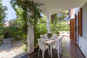 Une terrasse extérieure avec une table dressée pour le repas, entourée de chaises blanches, et protégée par une pergola couverte de plantes grimpantes.