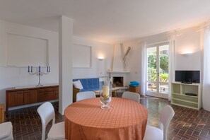 Un coin repas avec une table ronde, des chaises, un chandelier, une cheminée, un téléviseur et une vue sur la verdure à l’extérieur.