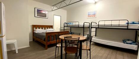 Una habitación compacta con una cama, literas, una mesa de comedor con sillas y un taburete blanco pequeño.