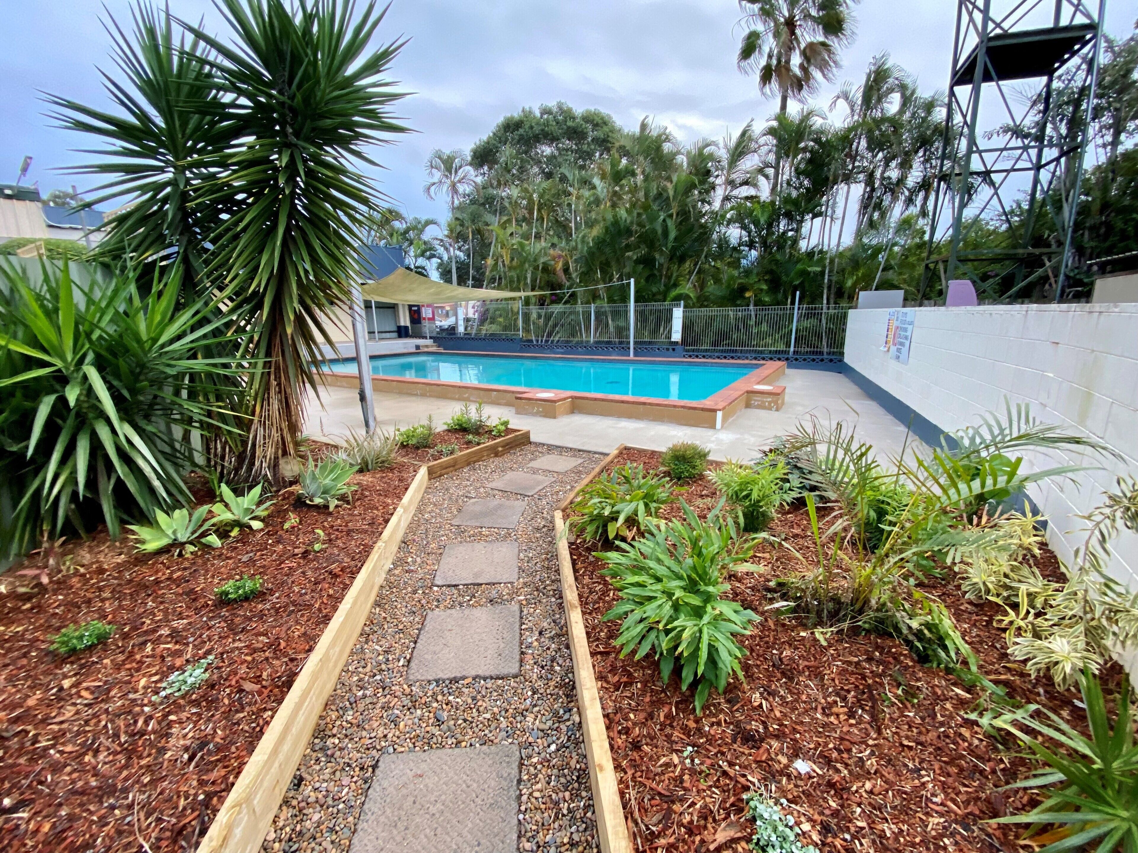 Zona de piscina con un sendero que lleva a ella, rodeada de plantas y un muro blanco.