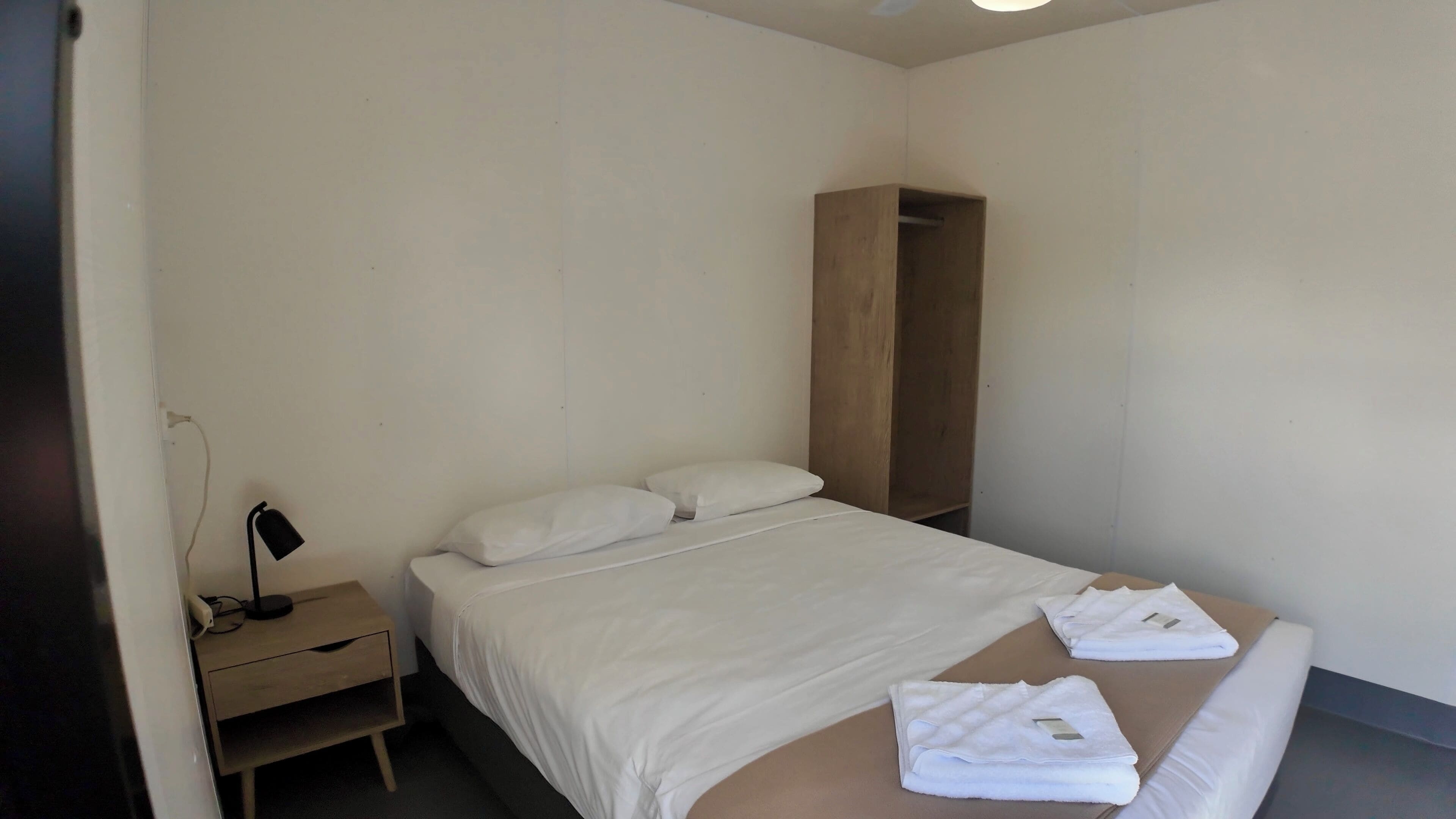 Een hotelkamer met een bed, nachtkastje, houten garderobe en bureau met lamp.