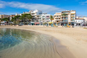 A beachfront hotel with multiple floors, a sandy beach, and palm trees. - Piso Claudia Beach Apartment with Terrace, Wi-Fi and Air Conditioning (Port de Pollença)