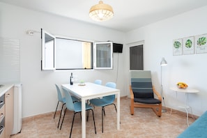 A room with a white table, blue chairs, a TV mounted on the wall, a small round table with a fruit bowl, and a chair with a striped cushion.