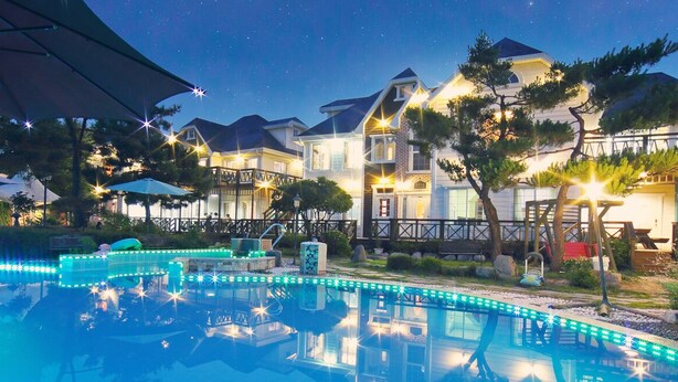 A swimming pool with blue LED lights, surrounded by a deck with umbrellas, and a row of houses with a starry night sky in the background. - Gyeongju Europe Village Pension (Gyeongju)