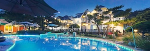 A swimming pool with blue LED lights, surrounded by a deck with umbrellas, and a row of houses with a starry night sky in the background.