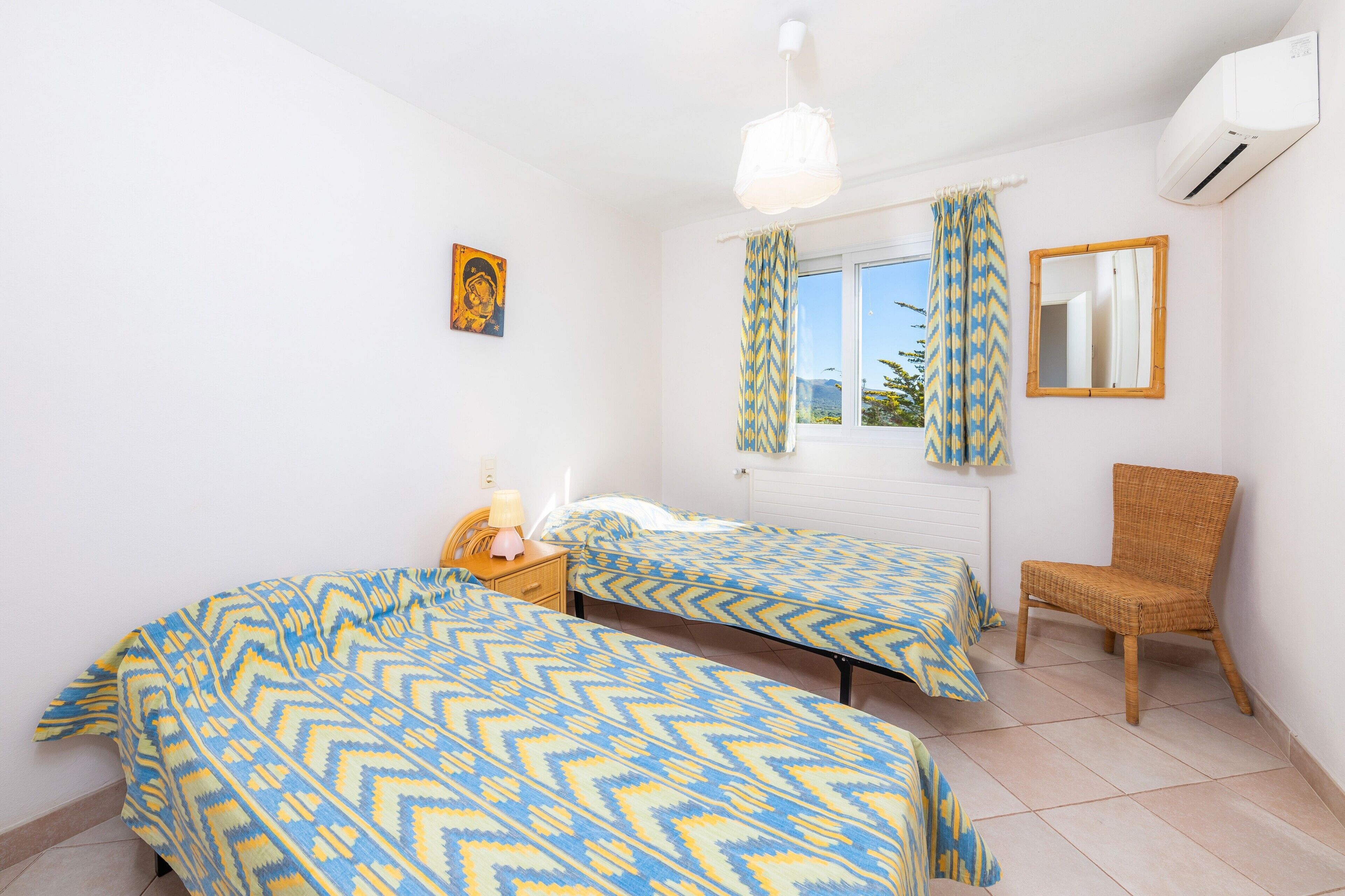 A bedroom with two beds, a chair, a window with curtains, a mirror, and an air conditioner.