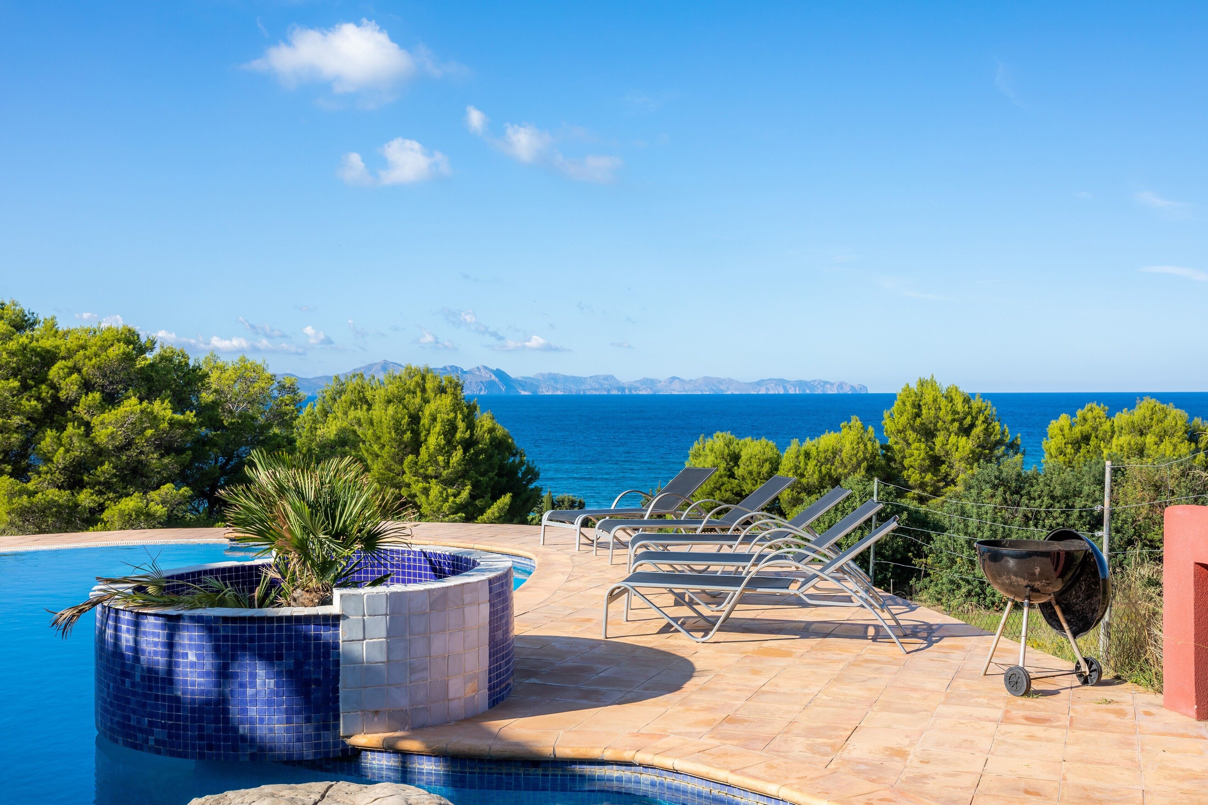 Un espace aménagé au bord de la piscine, avec des chaises longues, un jacuzzi et un barbecue, offrant une vue sur un plan d’eau et la verdure environnante.