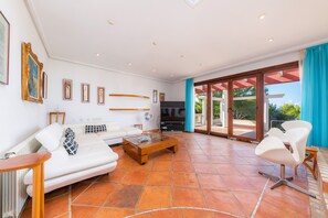 Un salón espacioso con un sofá blanco, una mesa de centro y un televisor. Se observan cuadros enmarcados en la pared y un amplio ventanal corredero que da a un área exterior.