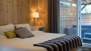 Ein schlichtes Schlafzimmer mit Holzwänden, einem Bett, Kissen, einem Nachttisch und einem Fenster mit Blick ins Freie.