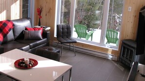 Ein gemütliches Wohnzimmer mit Ledersofa, grünem Sessel, Kamin und großem Fenster mit Blick auf Bäume.