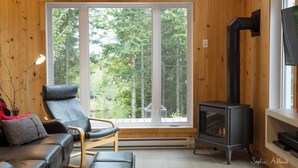 Ein gemütliches Zimmer mit einem großen Fenster, einem schwarzen Ledersessel, Holzboden und einem schwarzen Kaminofen.
