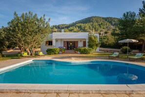 Una piscina con sillas de descanso, una casa con patio y un cerro cubierto de vegetación al fondo.