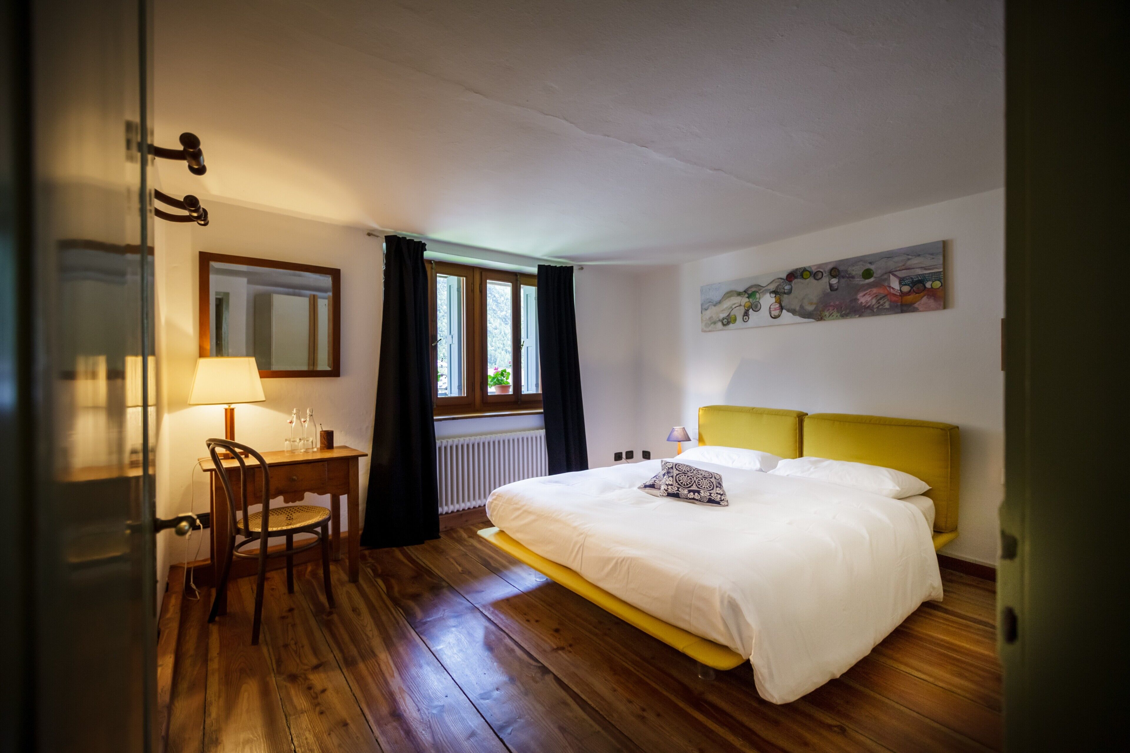 A bedroom with a bed, a desk, a chair, a window with curtains, and a painting on the wall.