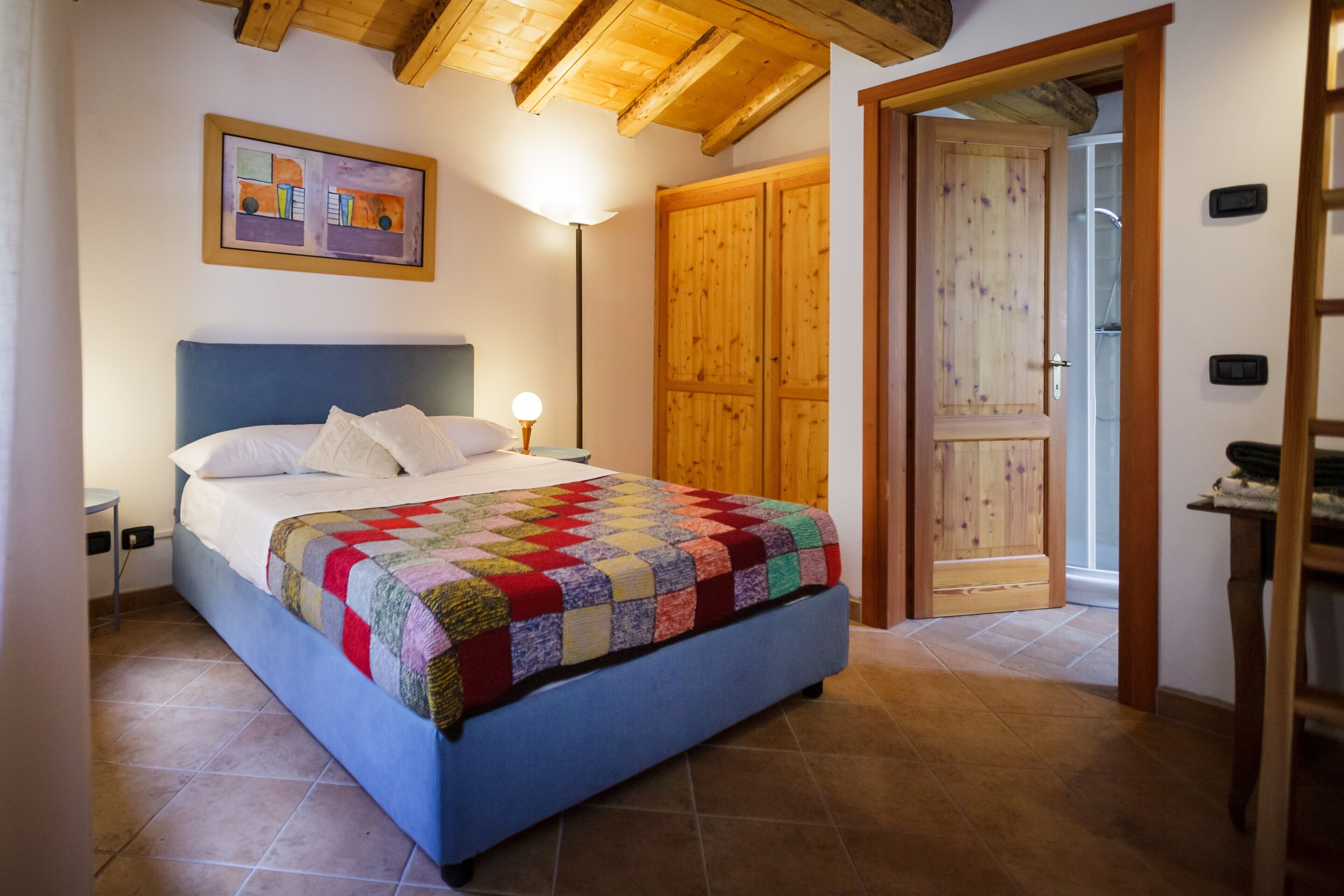 Une chambre avec un lit bleu, une couverture colorée, une porte en bois et un tableau accroché au mur.
