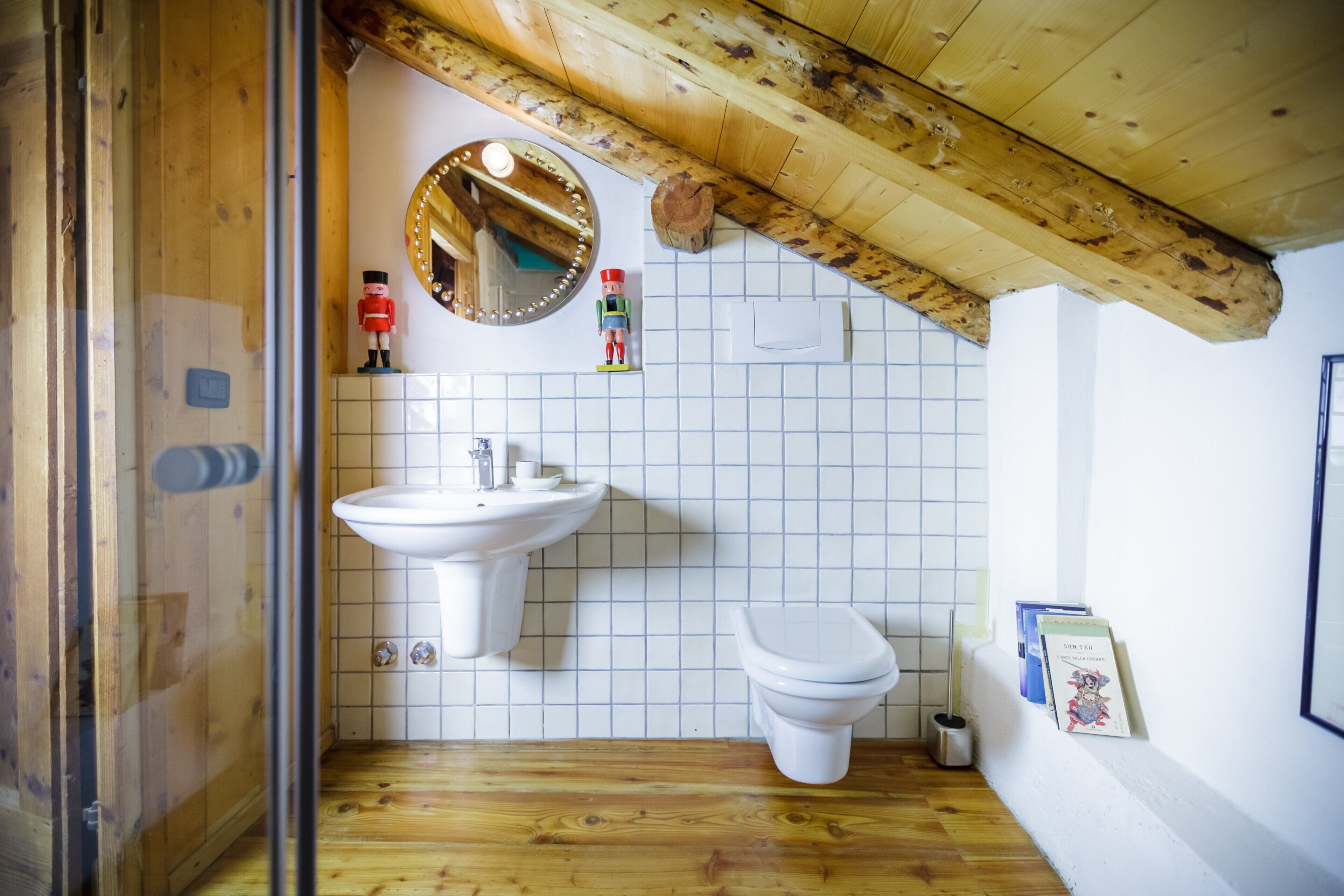 Ein Badezimmer mit Holzboden, weißen Fliesen, einem Waschbecken, einer Toilette, einem Spiegel und einer Holzzieldecke.