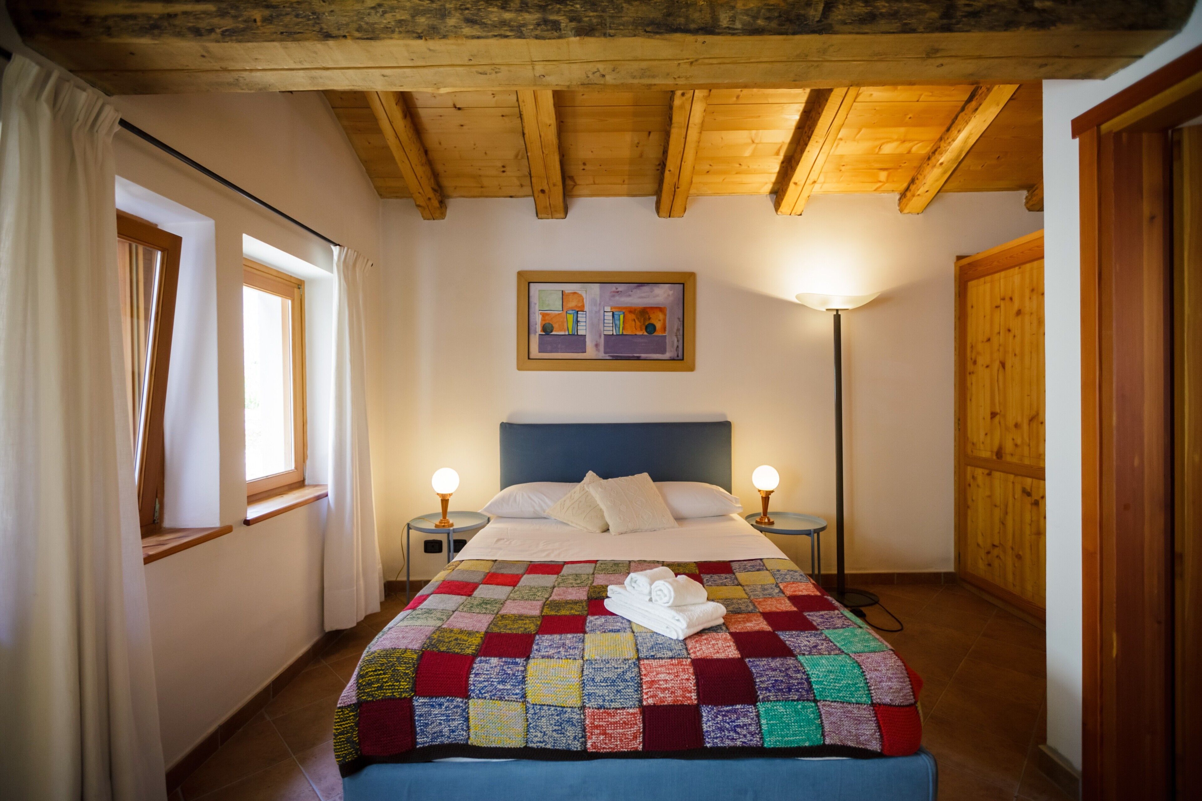 A bedroom with a wooden ceiling, a bed with a colorful quilt, two bedside lamps, and a painting on the wall.
