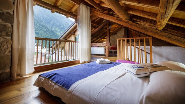 A bedroom with a wooden ceiling, a bed with white and blue bedding, a window with curtains, and a view of mountains.