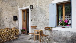 A rustic building with a wooden door, a window with shutters, a small table and chairs, and a stack of firewood.