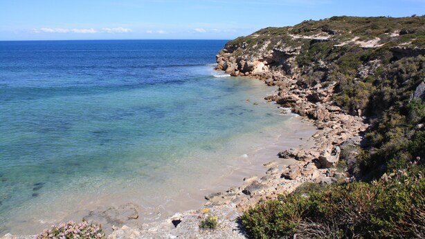 A coastal landscape with clear blue waters, rocky cliffs, and sparse vegetation.