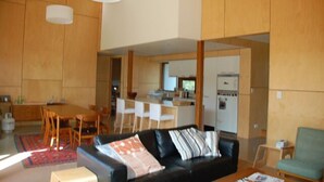 A modern living room with a black leather sofa, a dining area with wooden chairs, and a kitchen in the background. - The Architects House - Architecturally designed home in a scrubland setting with native birdlife and a bush track to the beach (Marion Bay)